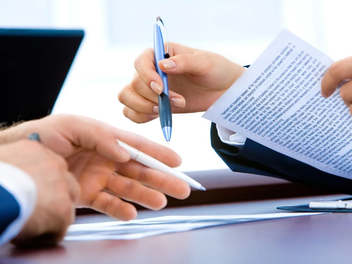 Two people seated at a desk reviewing a printed multi-page document, with one passing a pen across to sign it.