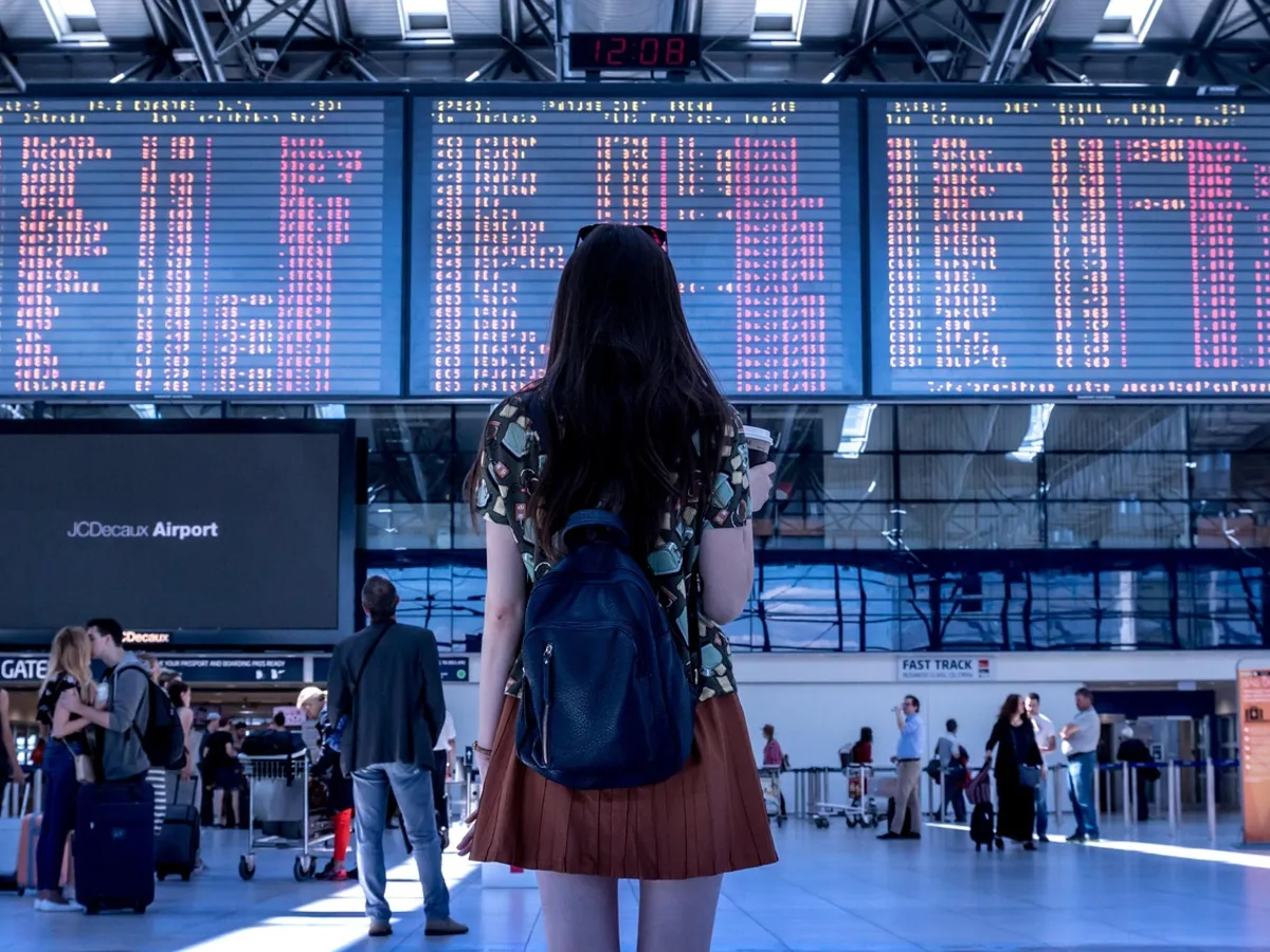 A young traveller with a backpack standing in a busy airport terminal, looking up at the illuminated departure boards.