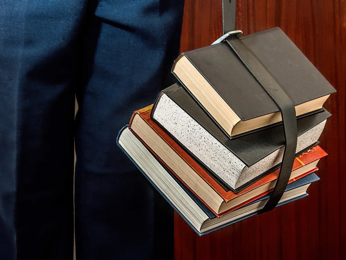 A person in a suit carrying a strapped stack of hardcover books at their side against a wood-panelled wall.