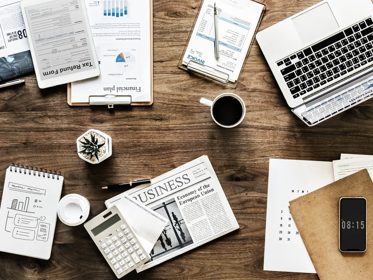 Overhead flat-lay of a wooden desk covered with paperwork and forms, a laptop, a calculator, a calendar, a coffee, and a phone showing the time.